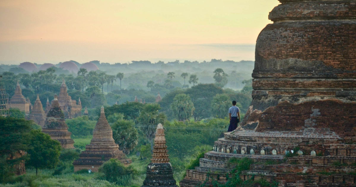 Traveller standing on ancient temple overlooking mist-covered pagodas across Southeast Asia at dawn
