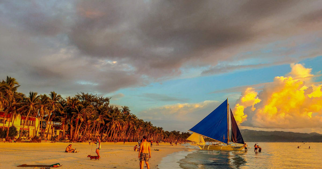 Tourists on Boracay beach at sunset with a traditional paraw sailboat in the Philippines